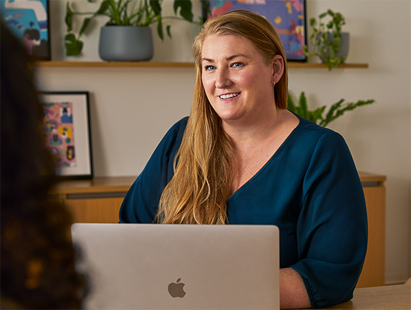 An Apple employee at a table in a common area, using a MacBook while speaking with a colleague.