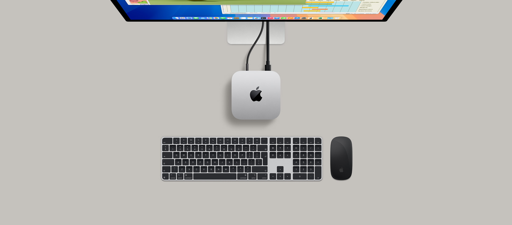 Top view of a silver Mac mini plugged into a display showing a game in active play, and next to a wireless keyboard, a gaming controller and blue AirPods Max