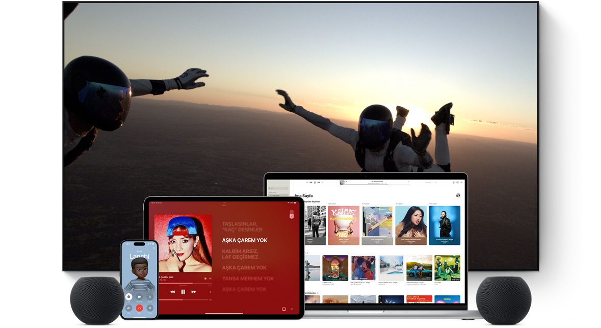 Geniş düz ekran bir televizyon gösteriliyor. Televizyonun önünde yan yana dizilmiş şekilde bir MacBook Pro, bir iPad, bir iPhone ve Gece Yarısı renginde bir HomePod mini yer alıyor.
