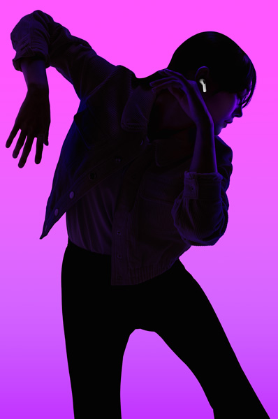 Silhouette of a person dancing with left arm angled down and right arm angled up, face is backlit in purple to highlight AirPods fitting securely in right ear.