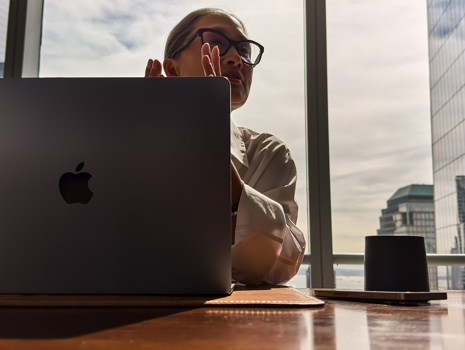 A woman sitting at a table in a highrise building. Behind her is a large window revealing other buildings. In front of her is an open MacBook.