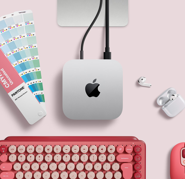 Top view of silver Mac mini in a desk setup with AirPods, a pink wireless keyboard, a pink wireless mouse, and a color swatch fan