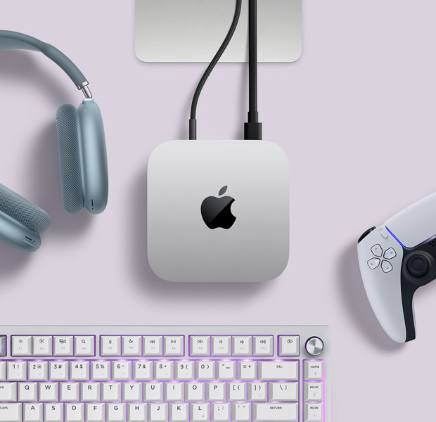 Top view of silver Mac mini, with two cords plugged into back ports, next to blue AirPods Max, a silver Magic Keyboard, and a wireless game controller